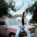 A young woman in casual attire sits on an old pink convertible car in an outdoor setting.