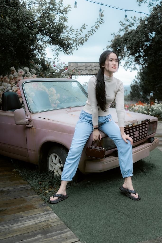 A young woman in casual attire sits on an old pink convertible car in an outdoor setting.