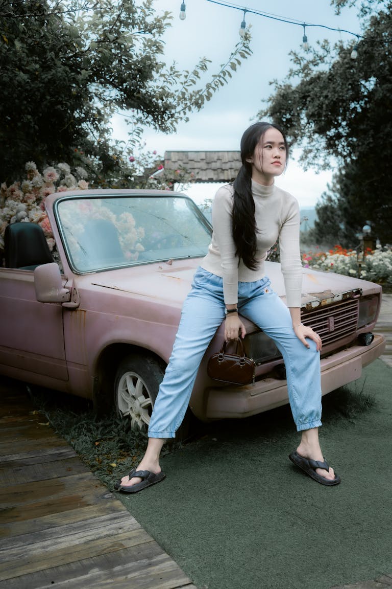 A young woman in casual attire sits on an old pink convertible car in an outdoor setting.